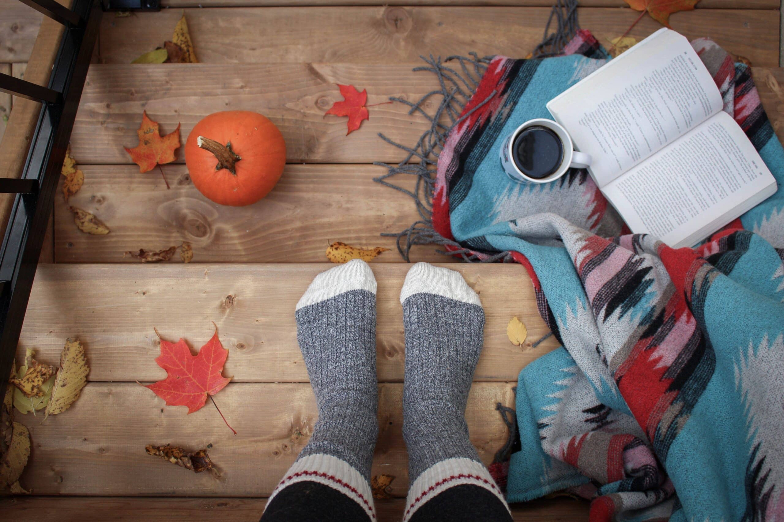 porch steps with view of feet with socks and fall foliage with a pumpkin, coffee and book