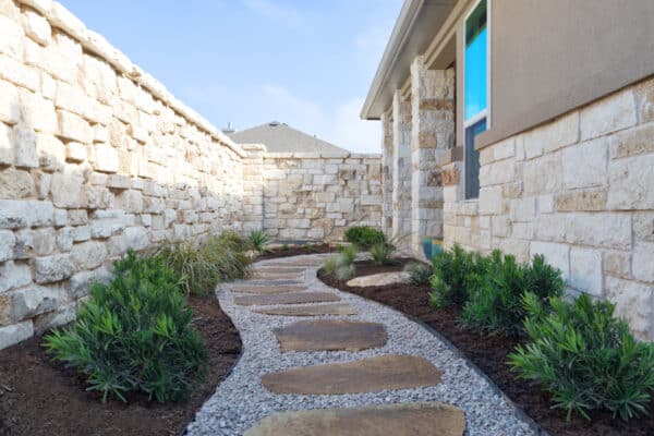 Side yard and back yard landscape design with gravel path and natural stone stepping stones with green plants along both sides along rock wall of backyard in central texas home