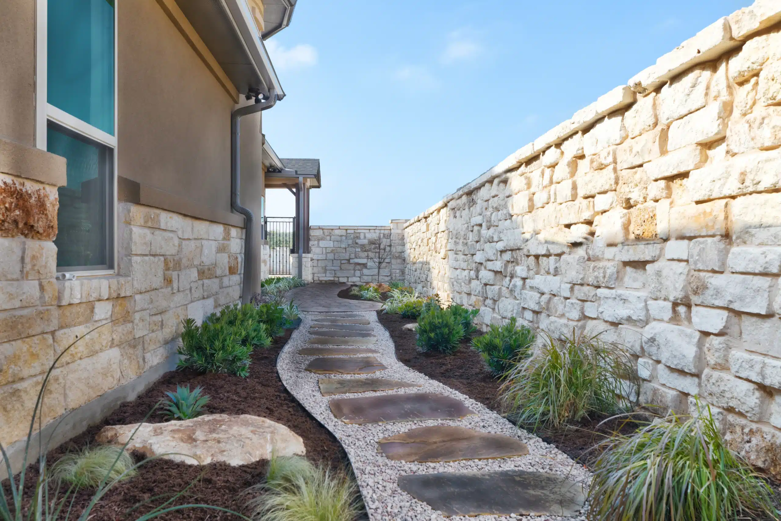 Natural stone walkway with gravel and steel edging alongside mulch plant beds with native plants and shrubs in the Austin area