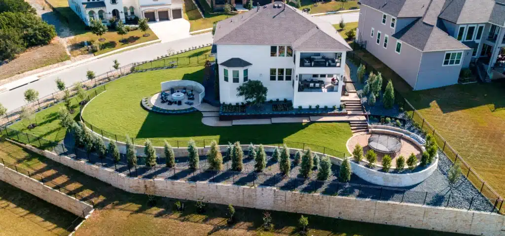 aerial view of backyard landscape design with gravel walkways, steel frame raised plant beds, circular seating area with fire pit, and circular rec area with trampoline, lined by spruce trees along lawn edges