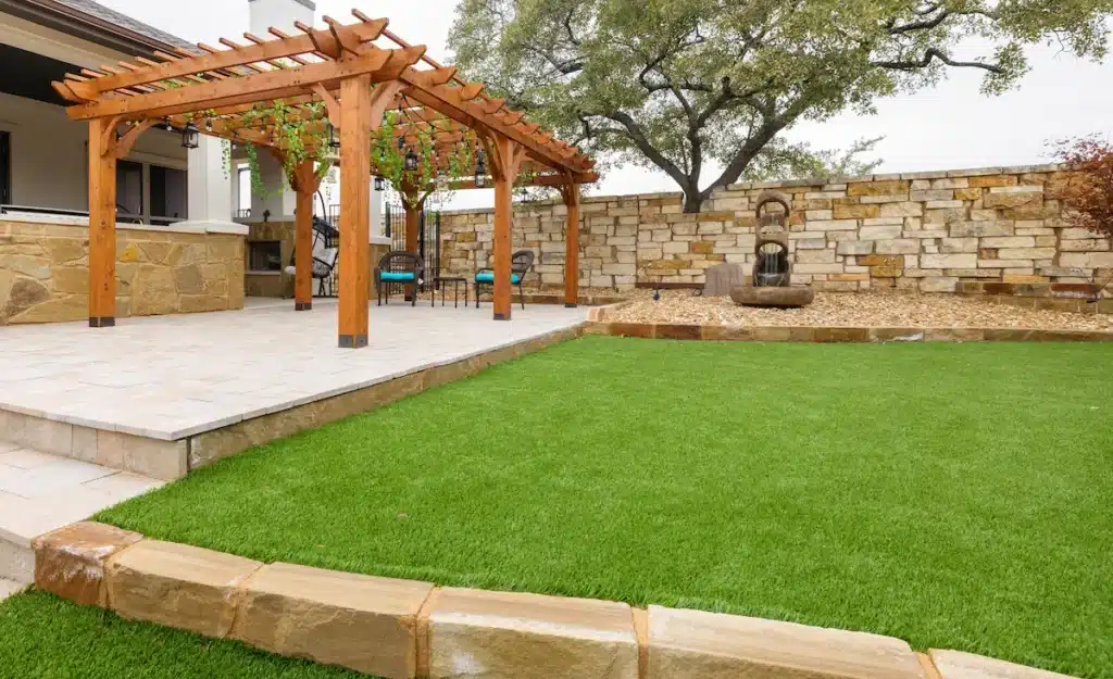 backyard with pergola on patio extension and grass lawn lined with rock wall behind outdoor water fountain sculpture